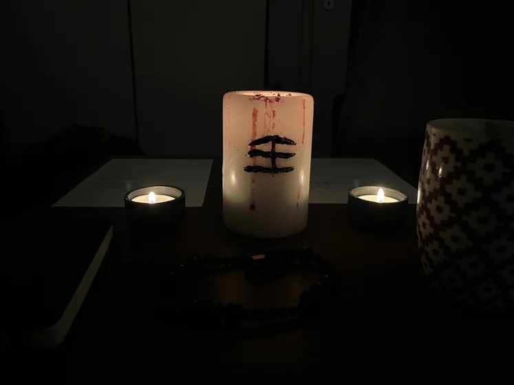 Ritual setup: Sacrifice book, prayer candles, the sacrifice candle in the center showing her holy sign, my prayer beads in front, and a cup of cocoa. Two pages written with a typewriter in the background.