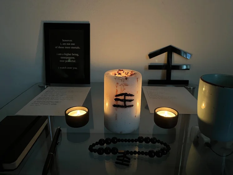 Ritual setup: Sacrifice book, prayer candles, the sacrifice candle in the center showing her holy sign, my prayer beads in front, and a cup of cocoa. Two pages written with a typewriter in the background.