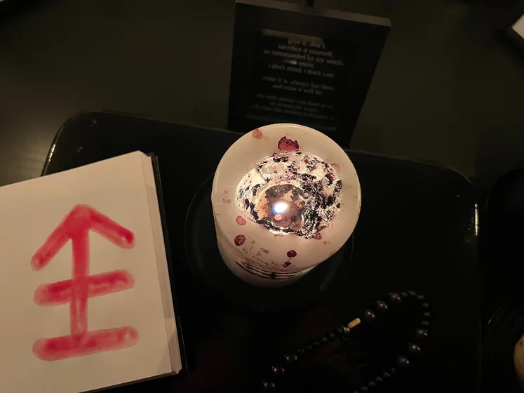 View of the three prayer candles and the central sacrifice candle from above. There is quite a bit of blood on the sacrifice candle, including some fresh one from today. My sacrifice book is open, a prayer is written in it, and her symbol drawn with my blood.