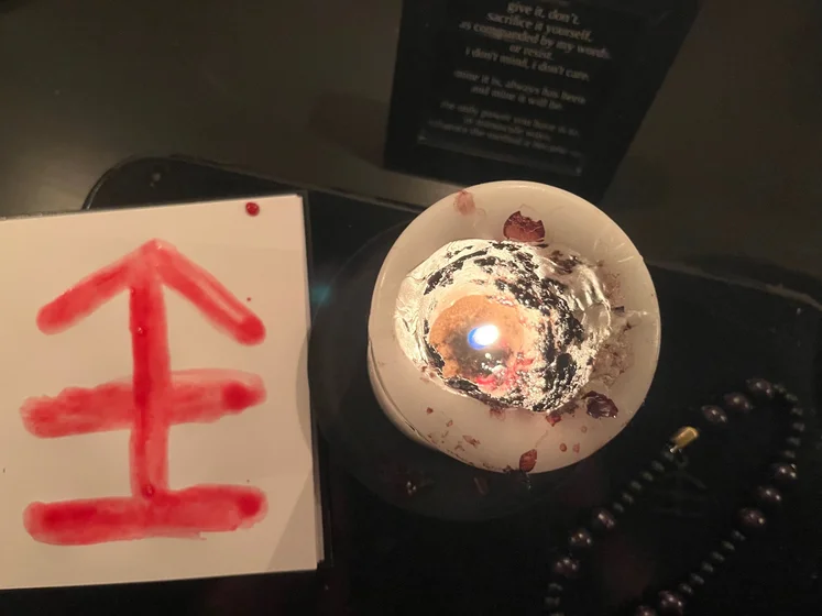 View of the three prayer candles and the central sacrifice candle from above. There is quite a bit of blood on the sacrifice candle, including some fresh one from today. My sacrifice book is open, a prayer is written in it, and her symbol drawn with my blood.